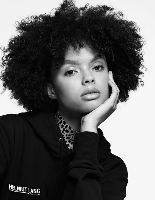 Portrait of a young Black woman with afro-textured hair, wearing a Helmut Lang hoodie in a black and white studio shot.