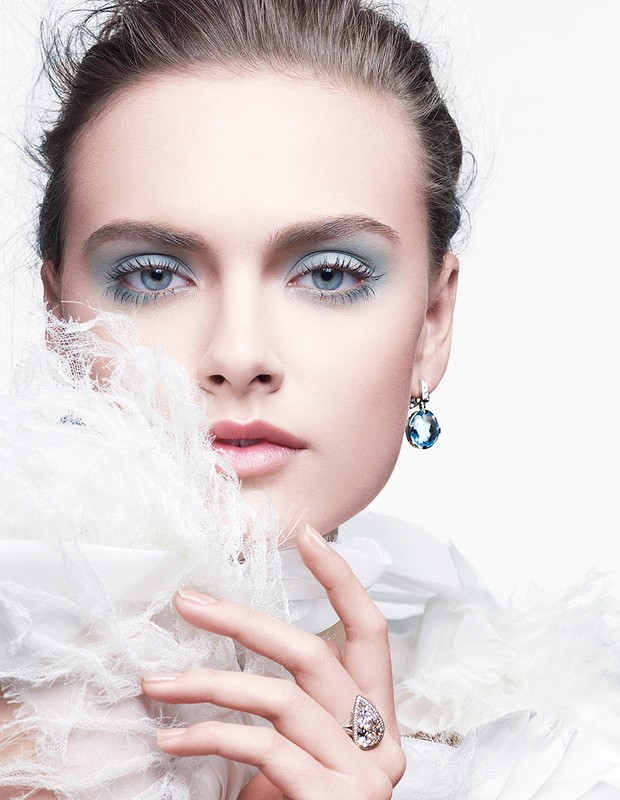 Close-up portrait of a woman with dramatic blue eye makeup, wearing a white feathered collar and sparkling jewelry against a white background.