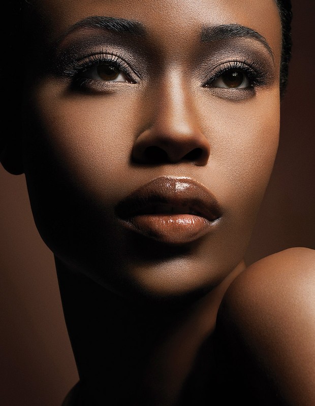 Close-up beauty shot of an African American woman with glossy brown lips, dark eyes, and smoky eyeshadow against a brown backdrop.