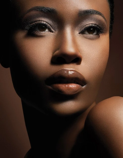 Close-up beauty shot of an African American woman with glossy brown lips, dark eyes, and smoky eyeshadow against a brown backdrop.
