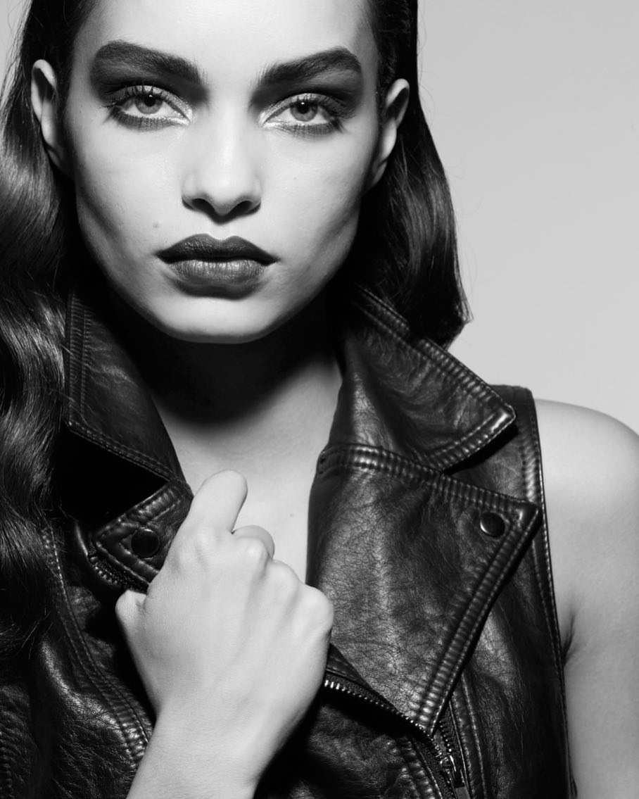 Black-and-white portrait of a woman with dark hair and dramatic makeup, wearing a leather vest and looking directly at the camera.