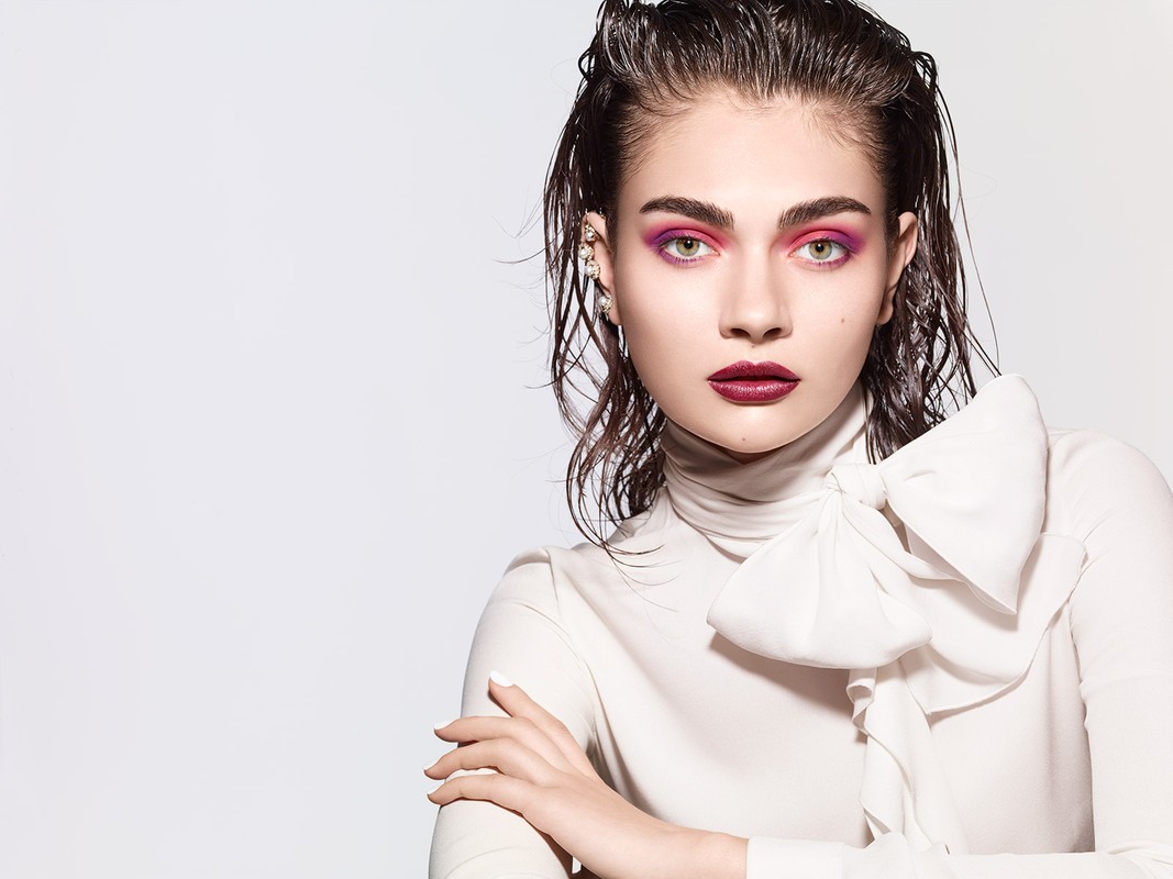 Close-up of a young woman with wet hair and bold makeup, posing in a white blouse with a large bow, set against a light background.