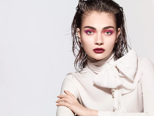 Close-up portrait of a woman with wet hair, vibrant pink eyeshadow, dark lipstick, and a white ruffled blouse against a plain background.