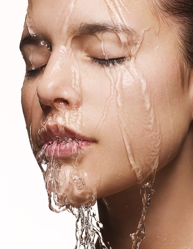 Close-up portrait of a woman with eyes closed as water cascades down her face against a clean white background, highlighting beauty.