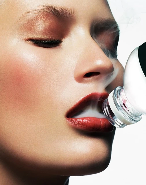 Closeup portrait of a fair-skinned woman with closed eyes, glossy red lips, and a beauty product bottle, with smoke or vapor effect.