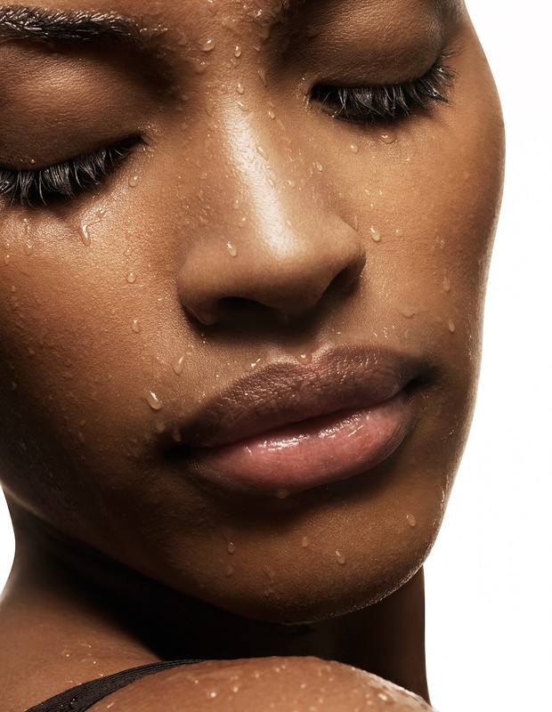 Close-up portrait of a Black woman with dark hair, wet skin glistening with water droplets against a plain white background.