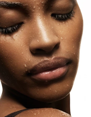 Close-up portrait of a Black woman with dark hair, wet skin glistening with water droplets against a plain white background.