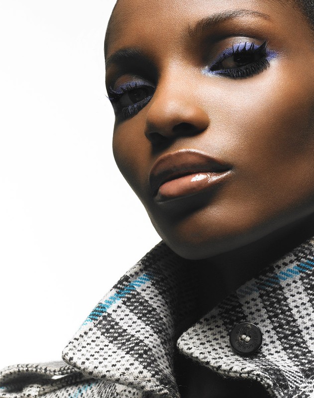 Close-up portrait of an African-American woman with bright blue eye makeup and a plaid coat against a white background in a studio.