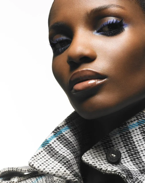 Close-up portrait of an African-American woman with bright blue eye makeup and a plaid coat against a white background in a studio.
