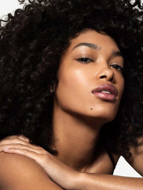 Close-up portrait of an adult woman with dark curly hair posing with her arms crossed in a photo studio setting.