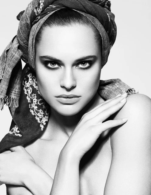 Striking black and white portrait of a woman with a patterned headscarf, intense gaze, and bare shoulders against a bright background.