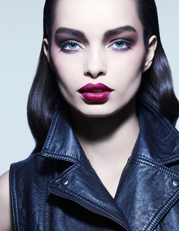 Close-up portrait of a woman with dark slicked-back hair wearing a black leather jacket against a light grey background.