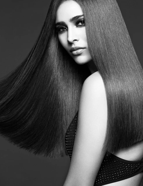 Monochrome portrait of a woman with long, straight hair and dark makeup, posing against a dark gray background in a beauty shot.