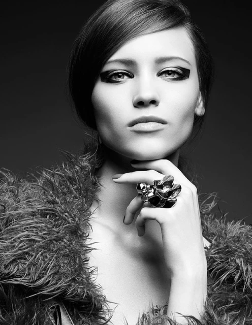 Black and white close-up studio portrait of a woman with dramatic eye makeup wearing an abstract statement ring and feathered collar.
