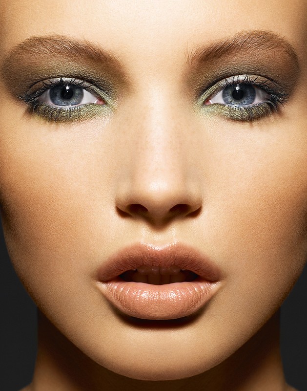 Close-up portrait of a woman with olive green eyeshadow and neutral lipstick against a dark grey background, highlighting beauty and makeup.