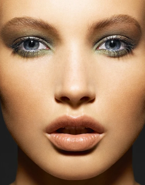 Close-up portrait of a woman with olive green eyeshadow and neutral lipstick against a dark grey background, highlighting beauty and makeup.