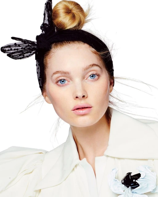 Close-up of a young fair-skinned woman with blue eyes, wearing a white jacket and a black lace headscarf, set against a white backdrop.