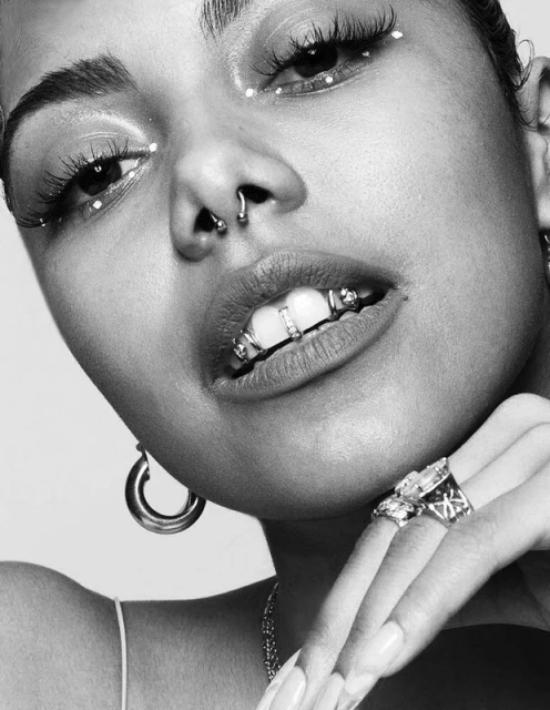 Close-up black and white portrait of a woman with septum piercing, ornate grill, heavy makeup, and jewelry posing in front of a plain background.