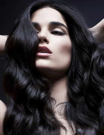 Close-up portrait of an adult woman with long, dark, wavy hair, a neutral facial expression, and fair skin, set against a dark background.