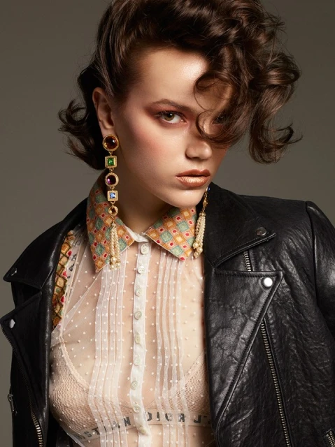 A woman in a sheer dotted blouse and black jacket looks at the camera, accessorized with statement earrings and gold lipstick, against a grey backdrop.