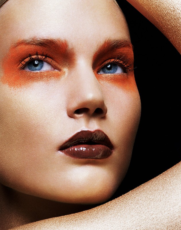 Close-up of a female model with bronze-toned skin, brown glossy lips, and bright orange makeup around the eyes on a black background.