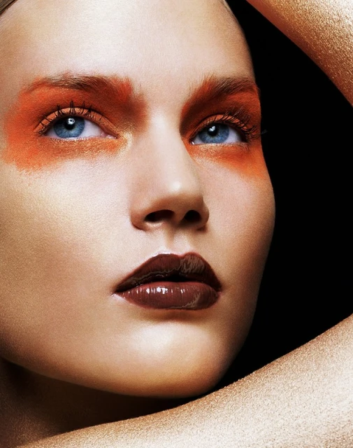 Close-up of a female model with bronze-toned skin, brown glossy lips, and bright orange makeup around the eyes on a black background.