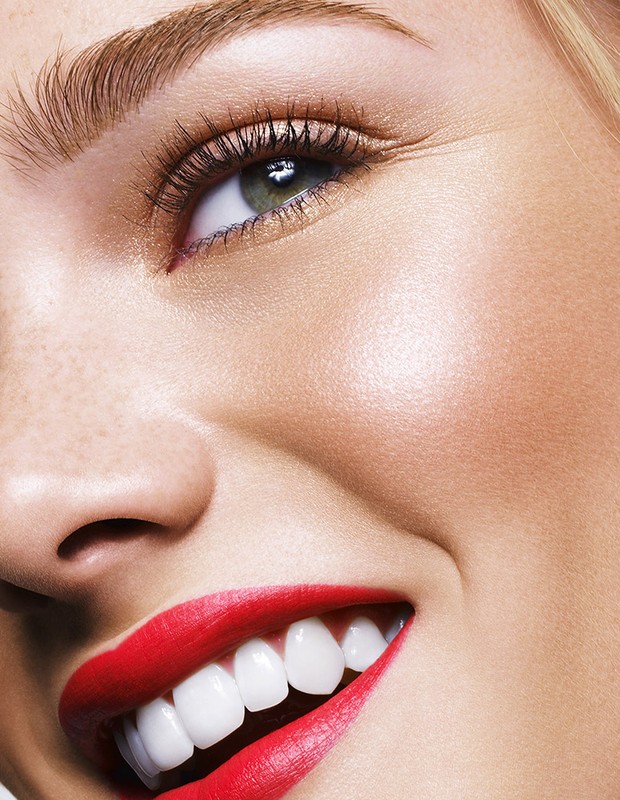Close-up of a smiling woman with bright red lipstick, showcasing her teeth, eye, and eyebrow. Focus is on beauty and facial details.