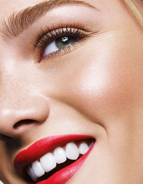 Close-up of a smiling woman with bright red lipstick, showcasing her teeth, eye, and eyebrow. Focus is on beauty and facial details.