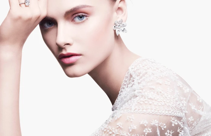 Close-up portrait of a woman wearing floral crystal earrings, a diamond ring, and a white lace top against a white background.