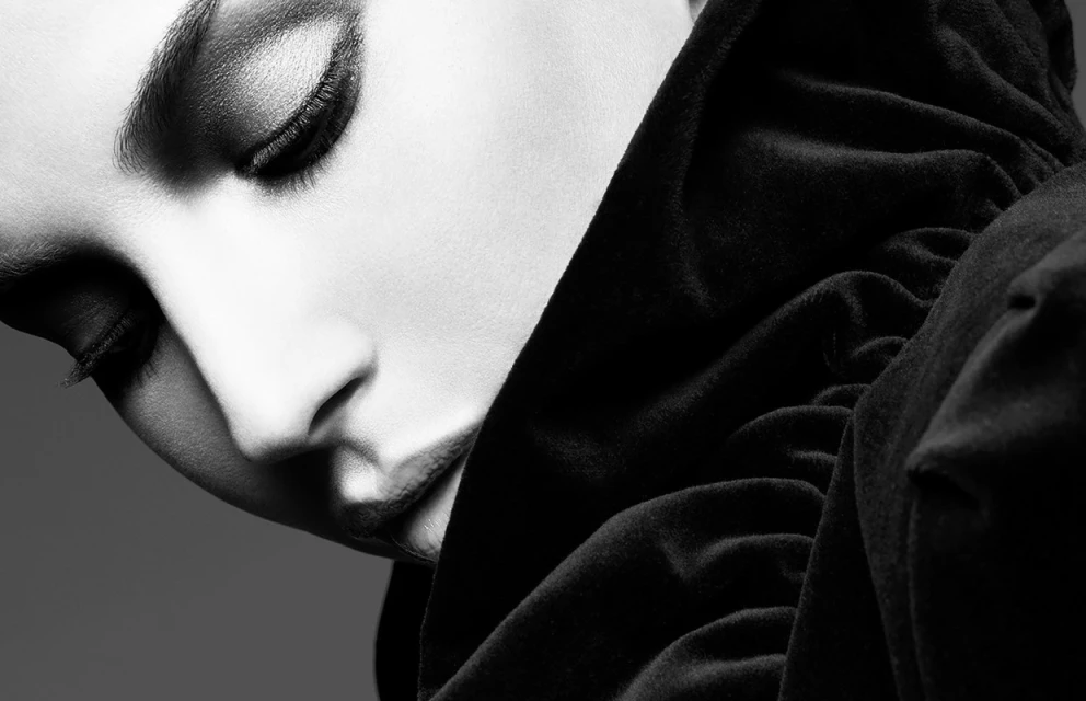 Close-up portrait of a woman with dramatic eye makeup wearing a dark, draped fabric, captured in a black and white studio setting.