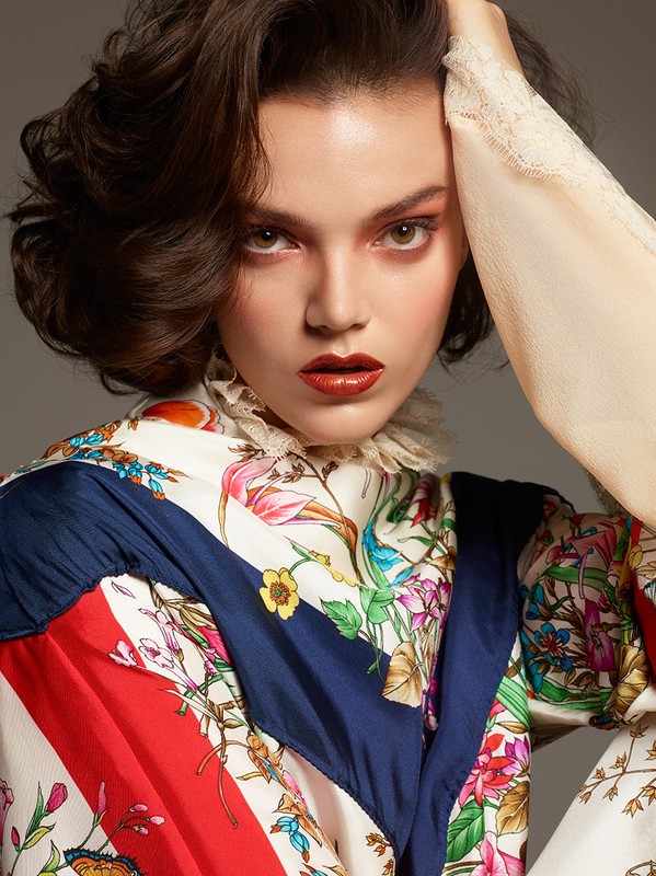 Close-up portrait of a woman with dark hair and red eye shadow, wearing a floral top against a solid gray background.