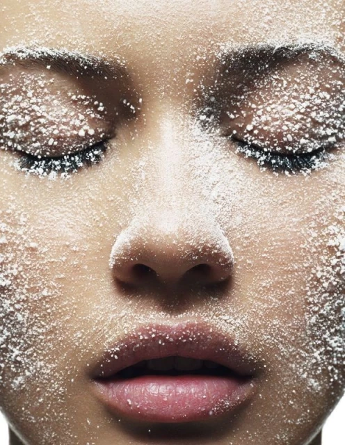 Close-up portrait of a woman's face covered in white powder, eyes closed, focusing on skin texture and makeup in a beauty context.