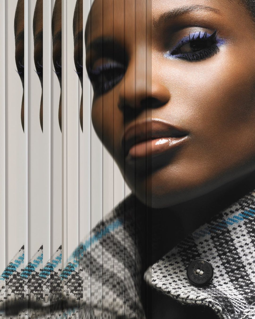 Close-up fashion shot of a black woman with blue eyeshadow, wearing a patterned coat, showcasing a modern editorial beauty aesthetic.