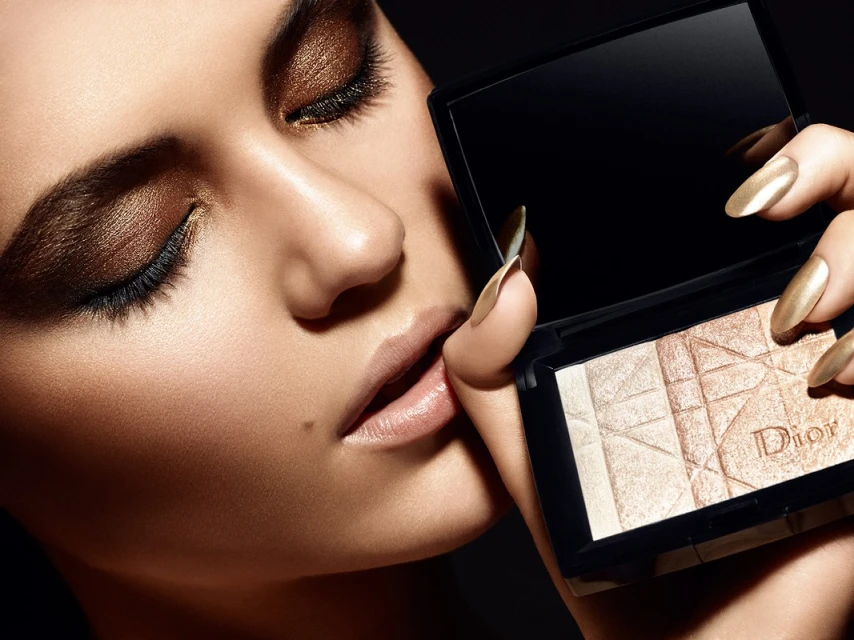 Closeup beauty shot of a woman with bronze eyeshadow, glossy lips, manicured gold nails, and holding a Dior makeup compact.