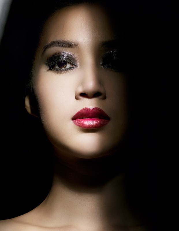 Close-up portrait of an Asian woman with dramatic makeup, including red lipstick and smoky eyes, set against a stark black background.