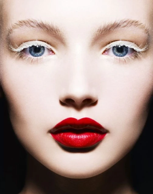 Extreme close-up of a young woman's face with bright red lipstick and artistic white eyelashes against a dark backdrop. Beauty shot.