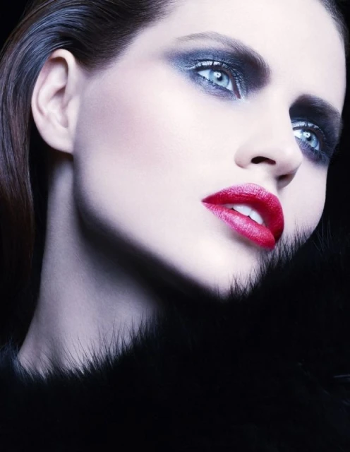 Woman with dramatic smokey eye makeup and red lipstick posing with a fur collar against a dark background in a beauty shot.