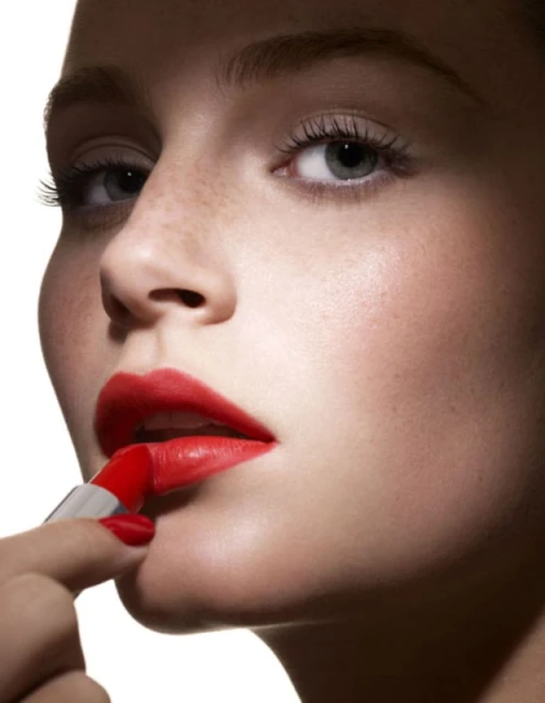 Close-up of a fair-skinned young woman applying bright red lipstick, showcasing cosmetic beauty and makeup artistry in a clean, bright setting.