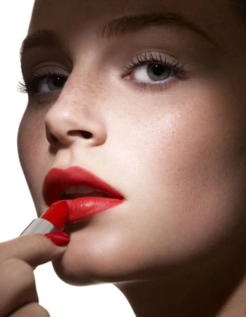Close-up of a fair-skinned young woman applying bright red lipstick, showcasing cosmetic beauty and makeup artistry in a clean, bright setting.