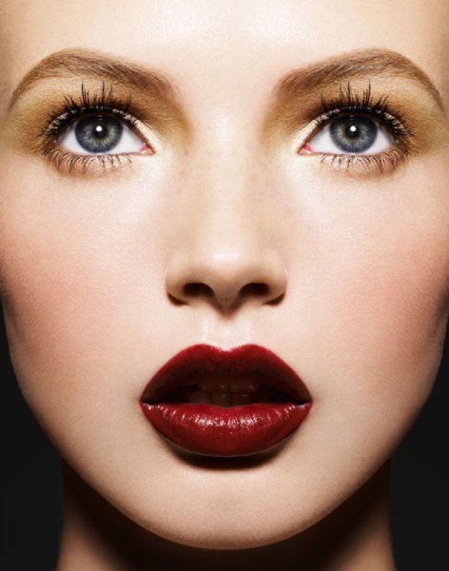 Close-up beauty shot of a fair-skinned young woman with blue eyes and deep red lipstick, eyes open, set against a dark gray background.