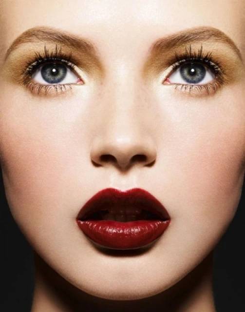 Close-up beauty shot of a fair-skinned young woman with blue eyes and deep red lipstick, eyes open, set against a dark gray background.