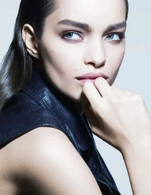 Close-up portrait of a young adult woman with a leather jacket, slicked-back hair, and fingers to her lips in a neutral studio setting.