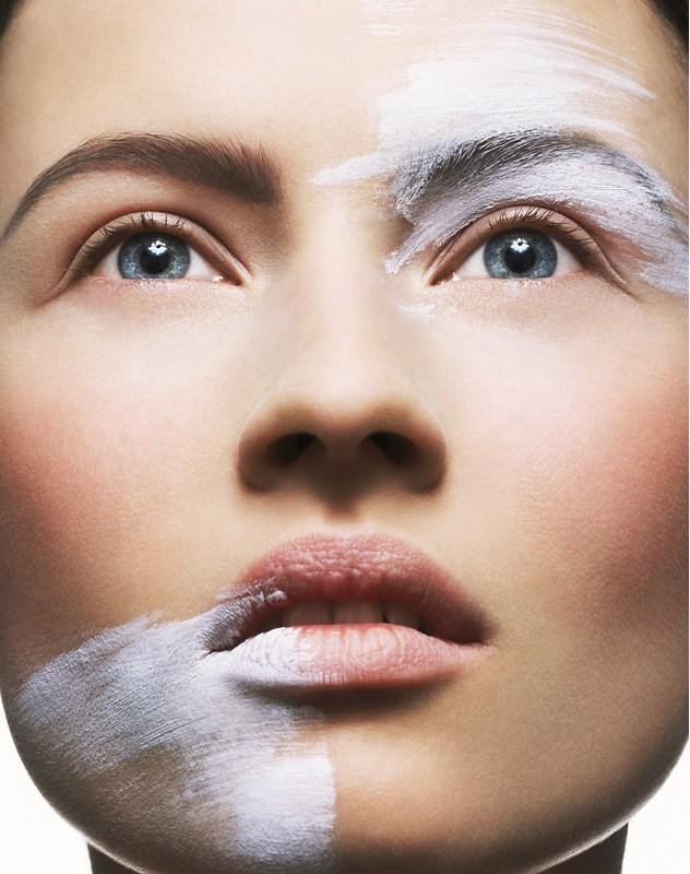 Close-up beauty shot of a woman's face, her features partially covered in white paint, emphasizing her blue eyes and facial symmetry.