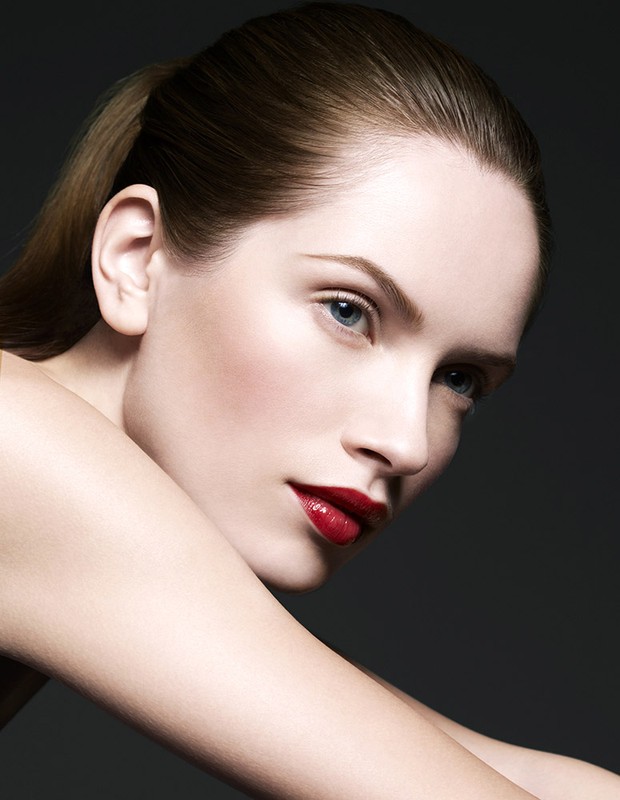 Close-up portrait of a fair-skinned woman with red lipstick and a ponytail, posing against a dark backdrop in a beauty-style shot.