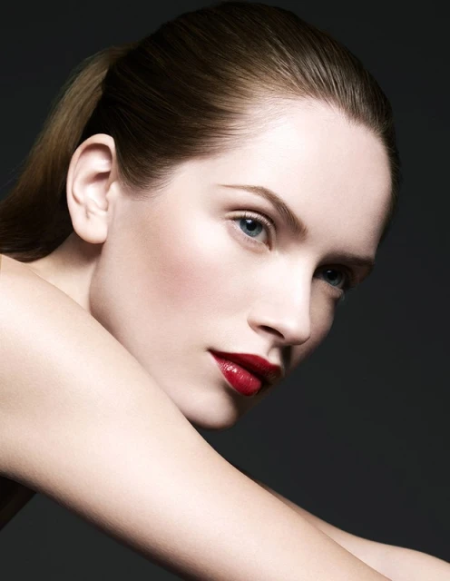 Close-up portrait of a fair-skinned woman with red lipstick and a ponytail, posing against a dark backdrop in a beauty-style shot.