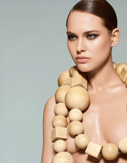 Close-up of a bare-shouldered young woman with wooden bead necklace, light eyeshadow, and slicked-back brown hair against a grey backdrop.
