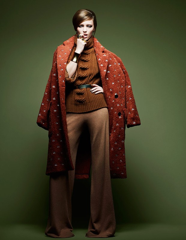 Model in rust patterned coat and brown turtleneck with flared trousers against olive backdrop by Philippe Salomon