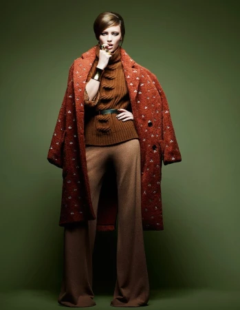 Model in rust patterned coat and brown turtleneck with flared trousers against olive backdrop by Philippe Salomon