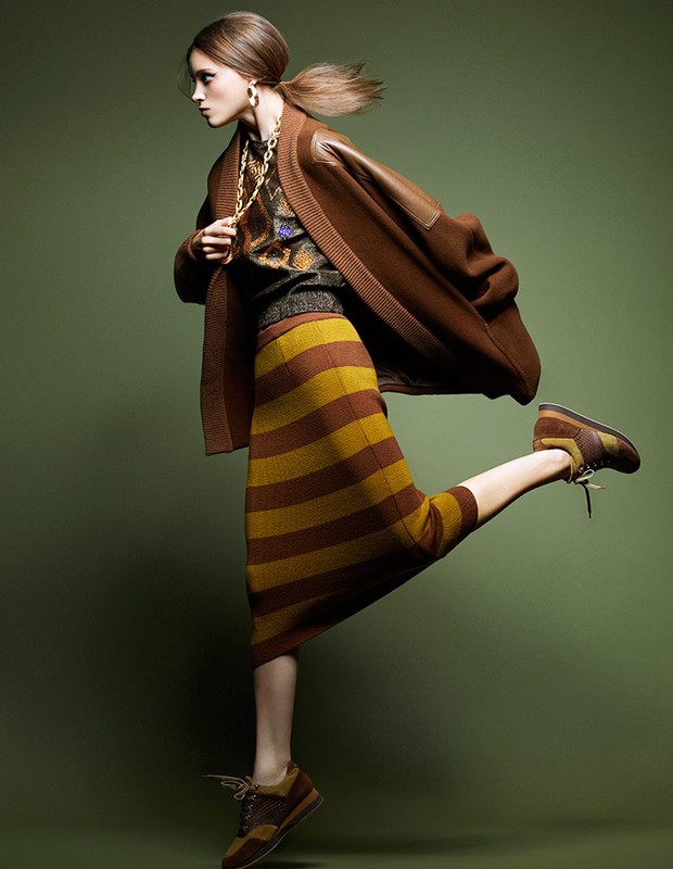A woman in a brown cardigan and striped skirt kicks one leg up in a fashion pose against a green background in a studio shot.
