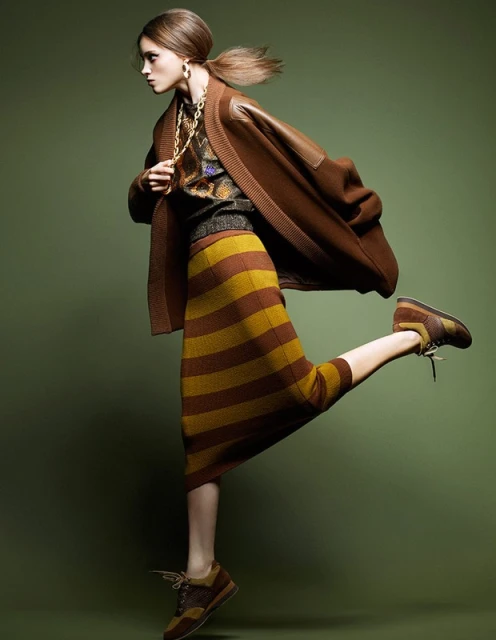 A woman in a brown cardigan and striped skirt kicks one leg up in a fashion pose against a green background in a studio shot.